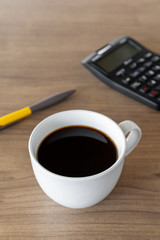hot coffee cup, pen and black calculator on wooden table with soft-focus and over light in the background