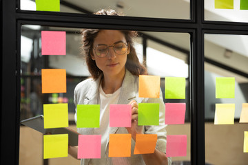Confident focused businesswoman wearing glasses writing ideas or tasks on sticky papers on glass...
