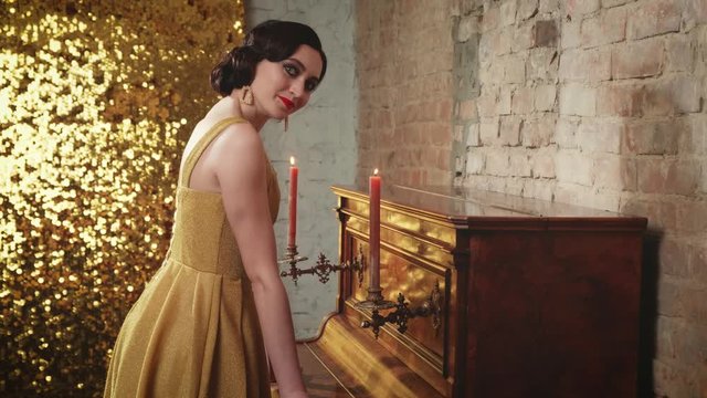 Beautiful young graduate woman. holiday evening bright makeup finger wave hairstyle. vintage luxury shiny golden dress. model posing near piano. backdrop brick wall. Christmas party retro fashion 1920