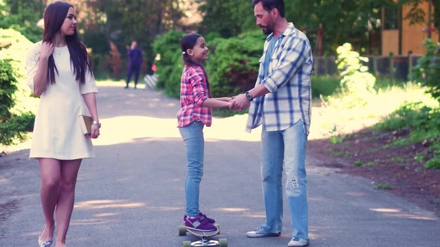 Father Holds Daughters Hands Halping Her To Stay Firm On Skateboard While Mother Walks Beside And Smile