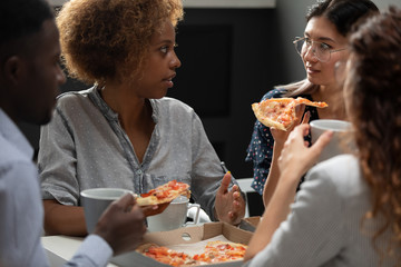 Diverse colleagues enjoying pizza together, African American and Asian businesswomen chatting during lunch break close up, employees team eating Italian fast food, talking, good relations