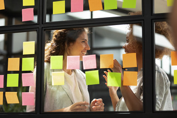 Happy diverse colleagues sharing creative startup ideas, working with sticky papers on glass wall...