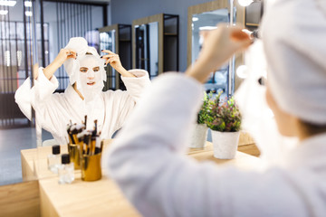 Young beautiful girl with a towel on her head and a white bathrobe cares for the face using a...