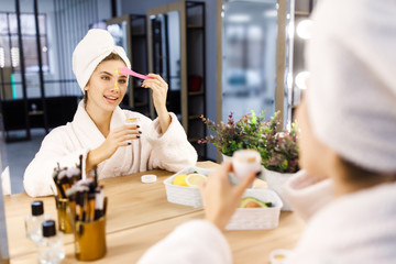 Young beautiful girl applying a gold mask to her face in front of a mirror.