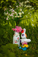 Easter decorative elements, vase with artificial flowers, watering pot with dandelions and bunny toy on green grass background.