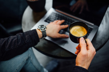 Cropped image, young businessman working on laptop, holding cup with coffee or latte with computer in modern cafe.