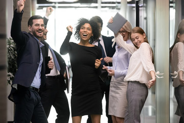 Overjoyed diverse businesspeople triumph have fun dancing in modern office corridor, happy multiracial colleagues coworkers celebrate business success together, engaged in Friday fun party at work - obrazy, fototapety, plakaty