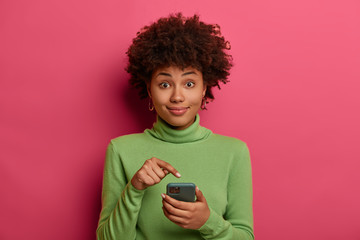 Afro American teenager points at modern smartphone display, recommends download new application,...