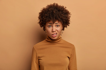 Fototapeta na wymiar Dissatisfied dark skinned young woman purses lips, looks unpleasantly at camera, expresses aversion, smirks face, wears clothes, isolated on beige background. Negative face expressions concept