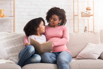 Young black pregnant mother reading book to her daughter at home