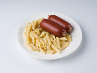 Sausages with a side dish of penne rigate. Food in a white plate on a light surface. Side view