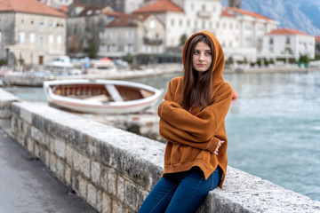 Fototapeta premium Woman in orange hoodie stands by the sea in city Perast on the background in Montenegro