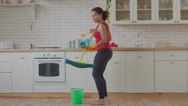 Joyful playful mixed race woman dancing and singing with mop while doing household chores in domestic kitchen. Excited pretty african american housewife with mop enjoying spring cleaning indoors.