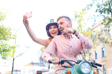 Happy young couple taking selfie on smartphone while sitting on scooter outdoors.