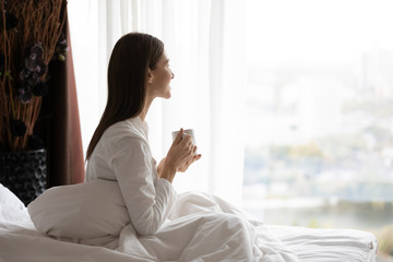 Obraz na płótnie Canvas Side view happy attractive young brunette woman sitting in bed under duvet, holding cup of black coffee, enjoying peaceful calm weekend vacation morning time alone in bedroom at home or hotel.