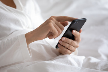 Close up focus on smartphone in female hands. Young woman in pajamas sitting in bed under duvet,...