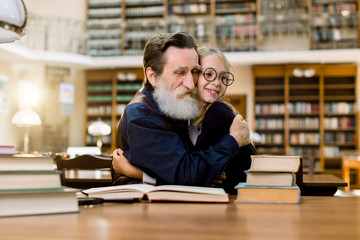 Grandfather and granddaughter or senoir teacher and pupil student, sitting at the table and hugging each other, in old vintage city library. Reading, education concept. Happy international book day