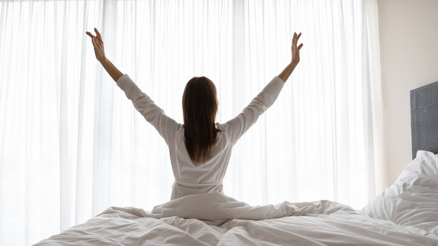 Back rear view young lady in night gown robe sitting on bed, rising arms up, stretching back after waking up. Energetic brunette woman looking at window, doing morning exercise at home or hotel.