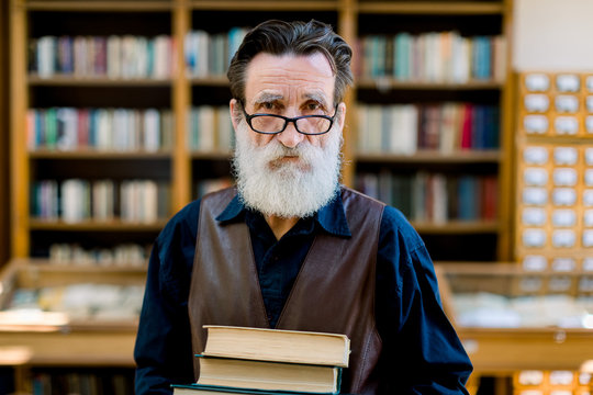 Close Up Portrait Of Handsome Bearded Senior Academic Professor Or Library Worker, Smiling And Holding Old Books, While Standing Over The Background Of Vintage Library Bookcases. Knowledge Concept