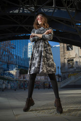 street fashion young stylish girl in a round black hat, round pink glasses and a dark dress posing against the backdrop of the city