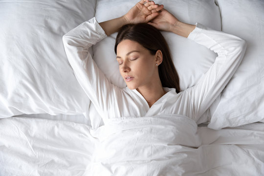 Top Above View Head Shot Serene Young Brunette Woman Lying On Soft Pillow With Raised Hands Under Duvet, Sleeping In Comfortable Bed, Enjoying Good Night Rest Alone In Hotel Or Home Bedroom.