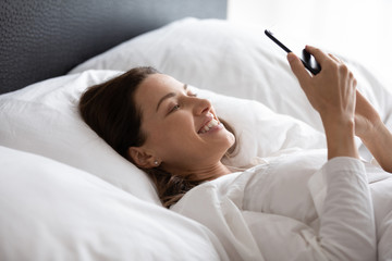 Obraz na płótnie Canvas Head shot close up side view happy smiling young woman relaxing in bed alone under blanket at home or hotel, looking at smartphone screen, reading sms with good news, feeling excited in morning.