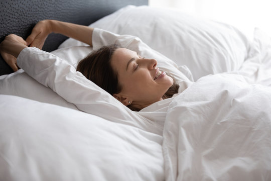 Head shot side view young happy woman lying in bed under duvet, raising arms, stretching back muscles, feeling refreshed in morning. Smiling brunette lady enjoying good holiday time after wake up.
