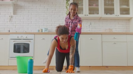 Carefree little mixed race daughter in rubber gloves with spray detergent piggybacking mother while housekeeping woman kneeling , washing kitchen floor using cleaning supplies during cleanup.