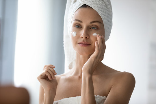 Head Shot Beautiful 30s Lady Looking In Mirror, Applying Hydrating Lotion Creme On Cheeks, Finishing Morning Domestic Skincare Routine. Smiling Woman Grooming Herself After Showering In Bathroom.