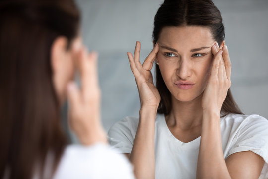 Head shot close up mirror reflection unhappy young brunette woman touching eyes area, feeling dissatisfied with first wrinkles, ineffective skincare products, confused with bad skin condition.