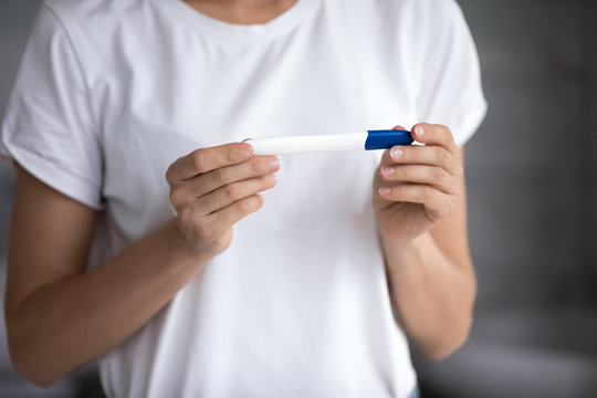 Cropped image young woman in white t-shirt holding quick pregnancy test, waiting for result. Focus on female hands with plastic stick, gynecologic healthcare, pregnancy confirmation or denial concept.