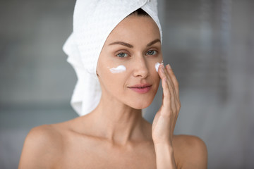 Peaceful attractive young lady with wrapped in towel head applying hydrating moisturizing creme, looking at camera. Beautiful pretty woman doing skincare routine after showering at home or hotel.
