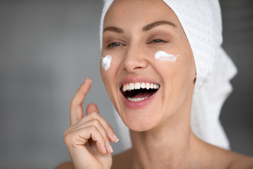 Overjoyed laughing young pretty woman with towel on head applying moisturizing creme on cheeks, feeling energetic after morning shower. Happy lady grooming herself in bathroom, skincare routine.