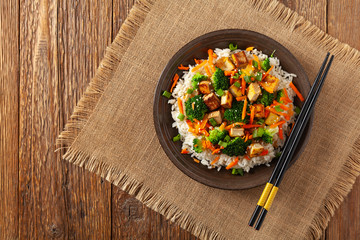 Tofu with rice and vegetables. Served on brown plate with chopsticks. Top view.
