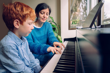 Piano teacher woman teaching a little boy at home piano lessons. Family lifestyle spending time together indoors. Children with musical virtue and artistic curiosity. Educational musical activities.