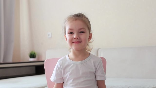 Four years little girl with pretty face smiling looking at camera at home sitting on chair near sofa, waving her head.