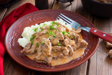 Traditional beef stroganoff in a ceramic bowl with mashed potato on a wooden table, selective focus