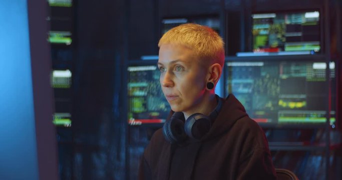 Female Caucasian woman siting in dark room of data center with many monitors and creating computer program. Woman hacking internet system of security.