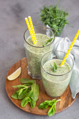 Green healthy smoothie in glasses with ingredients on wooden plate, grey stone background.