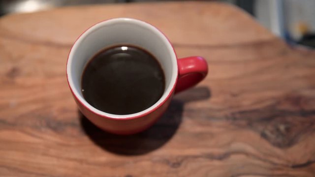 Close-up image of a red coffee cup on the outside and white on the inside resting on a wooden cutting board. A drizzle of steam rises from the cup. Blurry background.