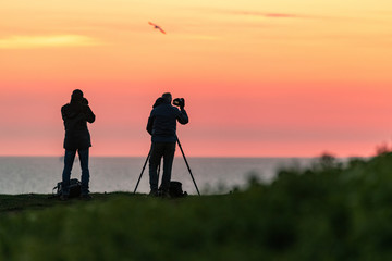 Silhouettes of two people standing on a cliff and photographing landscape and sea