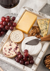 Glass of red wine with selection of various cheese in wooden box and grapes on light table background. Blue Stilton, Red Leicester and Brie Cheese with Cheddar and nuts with honey. Close up