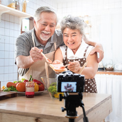 Couple senior Asian elder happy living in home kitchen. Grandfather wiping grandmother mouth after eating bread with jam vlog vdo for social blogger. Focus on camera. Modern lifestyle & relationship.