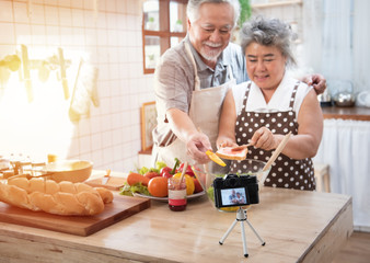 Couple senior Asian elder happy living in home kitchen. Grandfather and grandmother wiping bread with jam vlog vdo for social blogger. Focus on camera. Modern lifestyle & relationship.