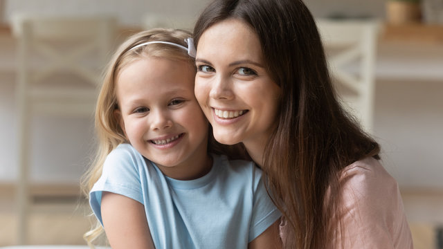 Close up portrait of smiling young mother and daughter cuddle hug look at camera posing for family picture, happy mom or nanny and small girl child embrace have fun show love and care