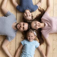 Top view of happy young parents and small children lying on warm wooden floor look at camera posing hold hands show unity and support, smiling family with kids demonstrate love and care