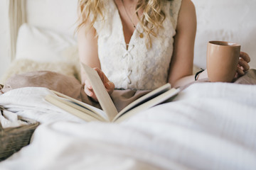 Closeup view of beautiful young blond woman sitting in her bed in bedroom in morning drinking hot...