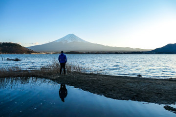 A man walking at the side of Kawaguchiko Lake and watching Mt Fuji, Japan. Reflection of the man in the water. Top of volcano covered with snow. Exploring new places. Soft sunset colors. Calmness
