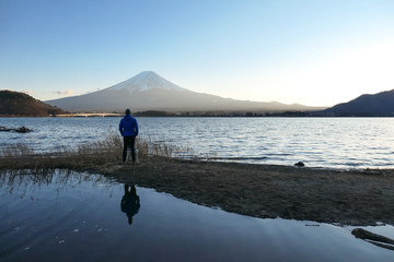 A man walking at the side of Kawaguchiko Lake and watching Mt Fuji, Japan. Reflection of the man in the water. Top of volcano covered with snow. Exploring new places. Soft sunset colors. Calmness