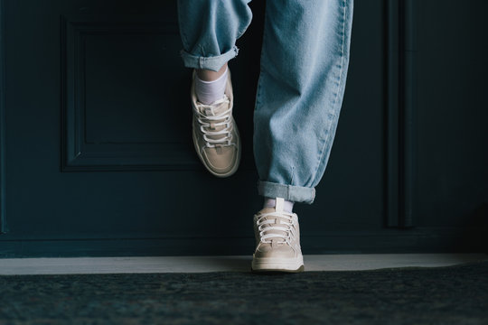 Closeup View Photography Of Two Female Legs Isolated At Black Wall Background. Woman Wearing Trendy Fashionable Blue Denim Jeans And Sneakers With Lacing.